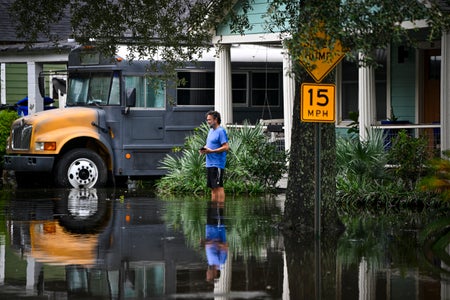 A man in blue T-shirt and shorts stands in front of his house in hi water with truck in background.