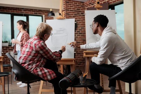 Art teacher supervising student explaining sketching technique while pointing to the student's drawing of a vase on their easel in a classroom